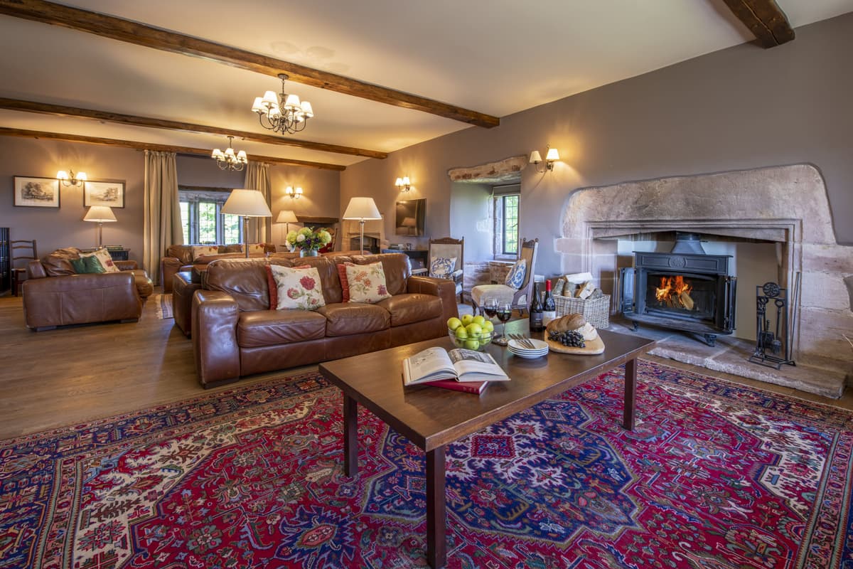 A beamed living room with a large patterned rug, leather sofas facing a lit wood burner in a stone fireplace, and lamps on