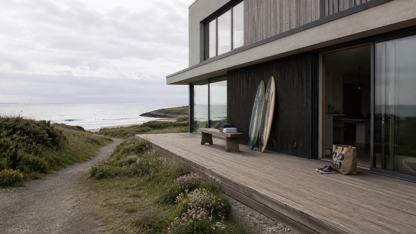 A modern UK coastal beach house with pale render and charred timber cladding, large picture windows facing the sea, and a wide wooden deck. Surfboards lean against the wall, a jute tote and canvas plimsolls sit beside an open sliding door. Wild beach grasses and sea thrift around the property, a path leading down to a sandy beach in the soft-focus background. Bright overcast UK coastal afternoon.