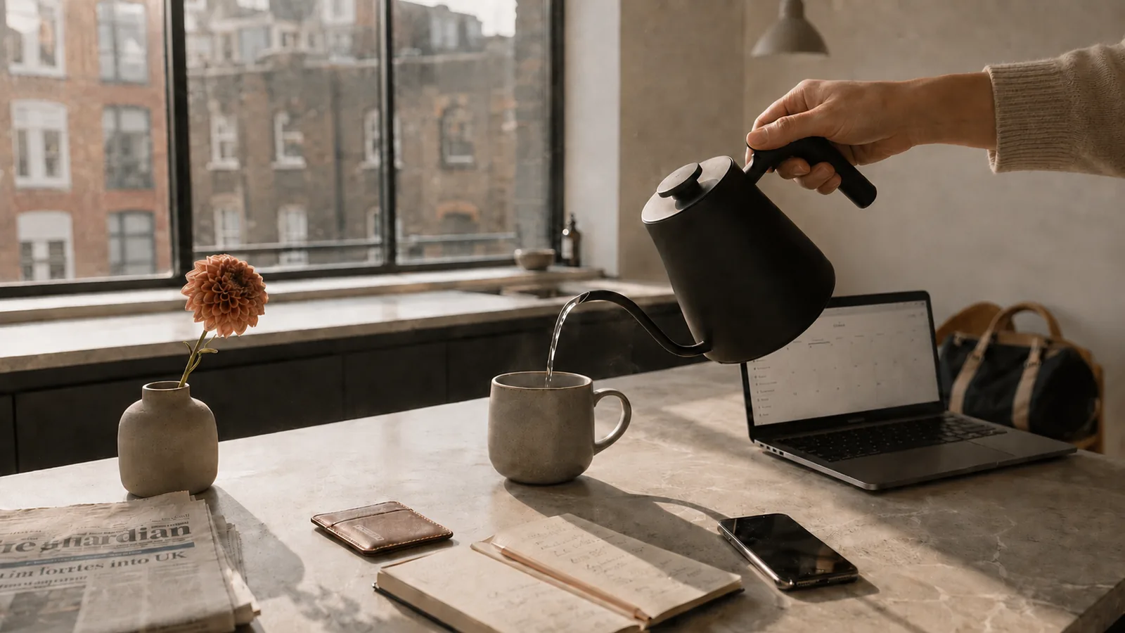 A modern UK city flat kitchen: a hand pouring water from a matte-black gooseneck kettle into a stoneware mug, beside a laptop showing a calendar, a notebook with handwritten plans, a folded newspaper, and a single dahlia in a small ceramic vase. Sunlight streaming through a slim-frame metal window with a view of London brick buildings.