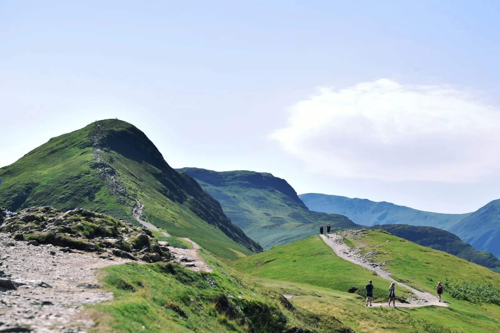 Walkers on a grassy ridge path heading toward a green fell summit in the Lake District, with more fells rolling into the distance under a bright summer sky